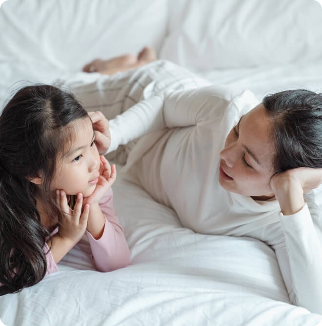 A woman and a young girl lying on a bed, facing each other and smiling, sharing a tender moment.
